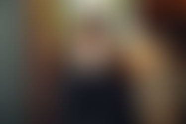 A person in black covers their face with hands, sitting on a chevron-patterned chair. Plants and a bright interior are in the background.