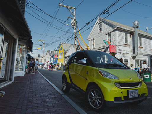 Bright green car on a busy street lined with shops. People walking, chatting. Blue sky, colorful storefronts create a lively atmosphere.