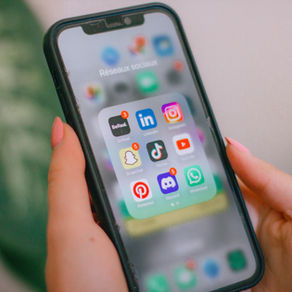 Hands holding a smartphone showing a French social media folder with BeReal, LinkedIn, Instagram, Snapchat, TikTok, YouTube and WhatsApp icons.
