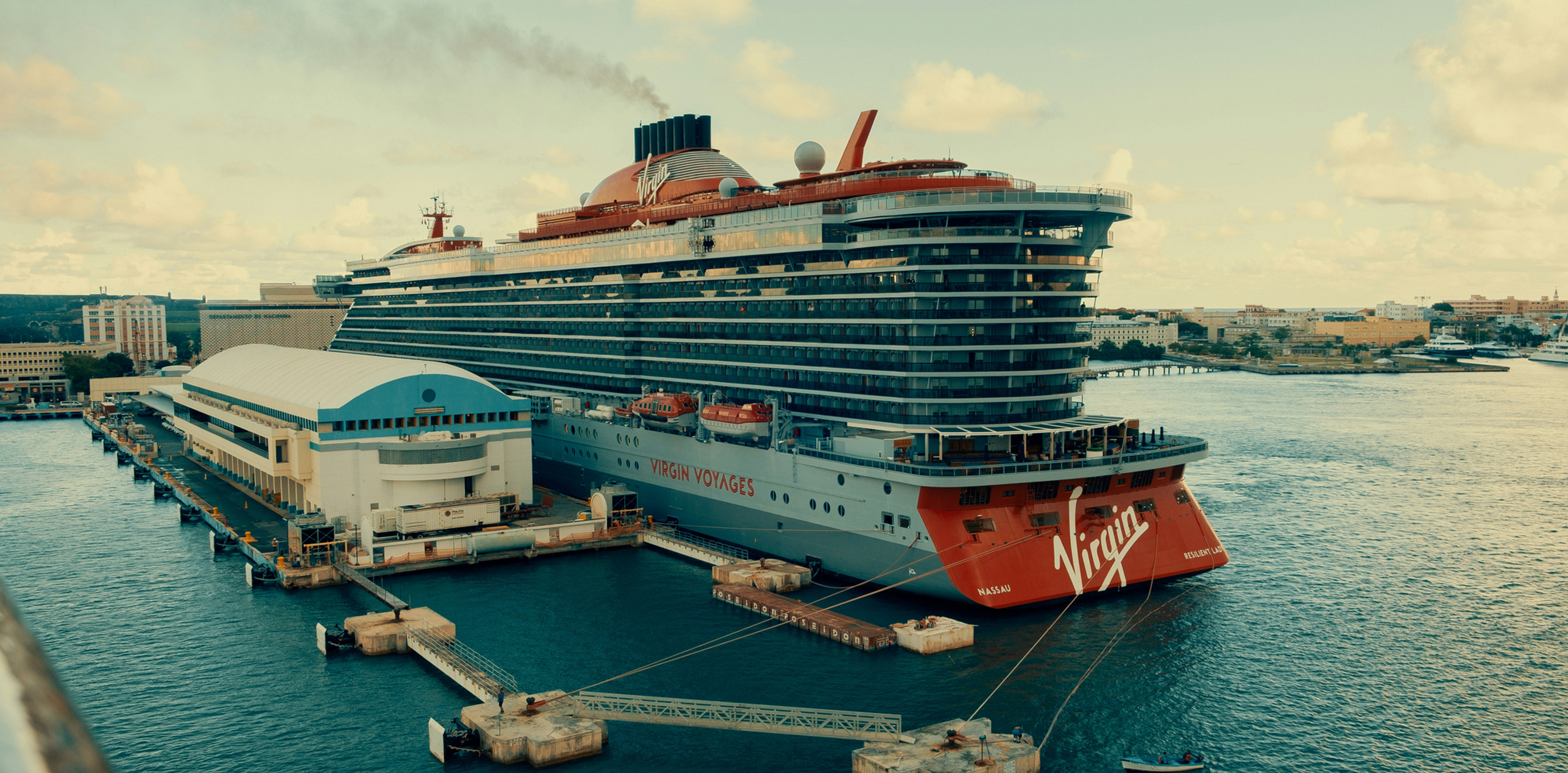 Large Virgin Voyages cruise ship docked at a harbor, with blue water, port buildings, and a cloudy sky at sunset