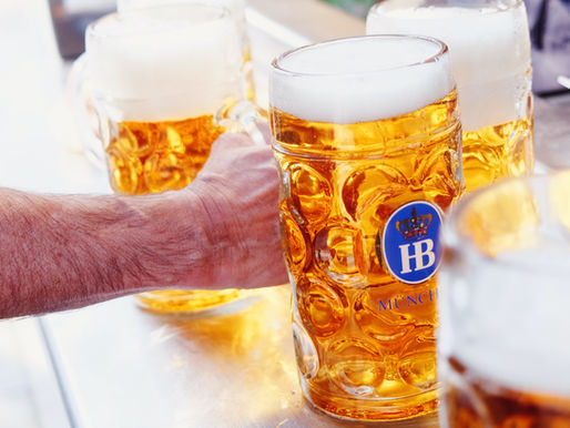 Hand holding a frosty HB Munch beer mug among several foamy amber beers on a metal table, bright pub scene