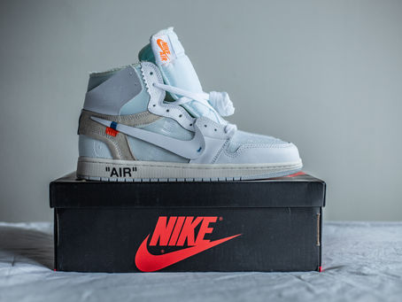 White sneaker on a black Nike box with red logo. "AIR" text on sole. Gray background, minimalistic setting. Sneaker has orange tag.