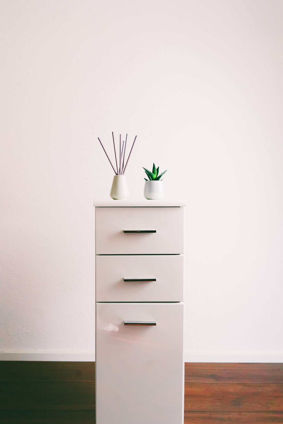 image d'une petite commode blanche minimaliste sur du parquet brun avec fond blanc pour illustrer un petit espace de rangement dédié pour un nouvel arrivant