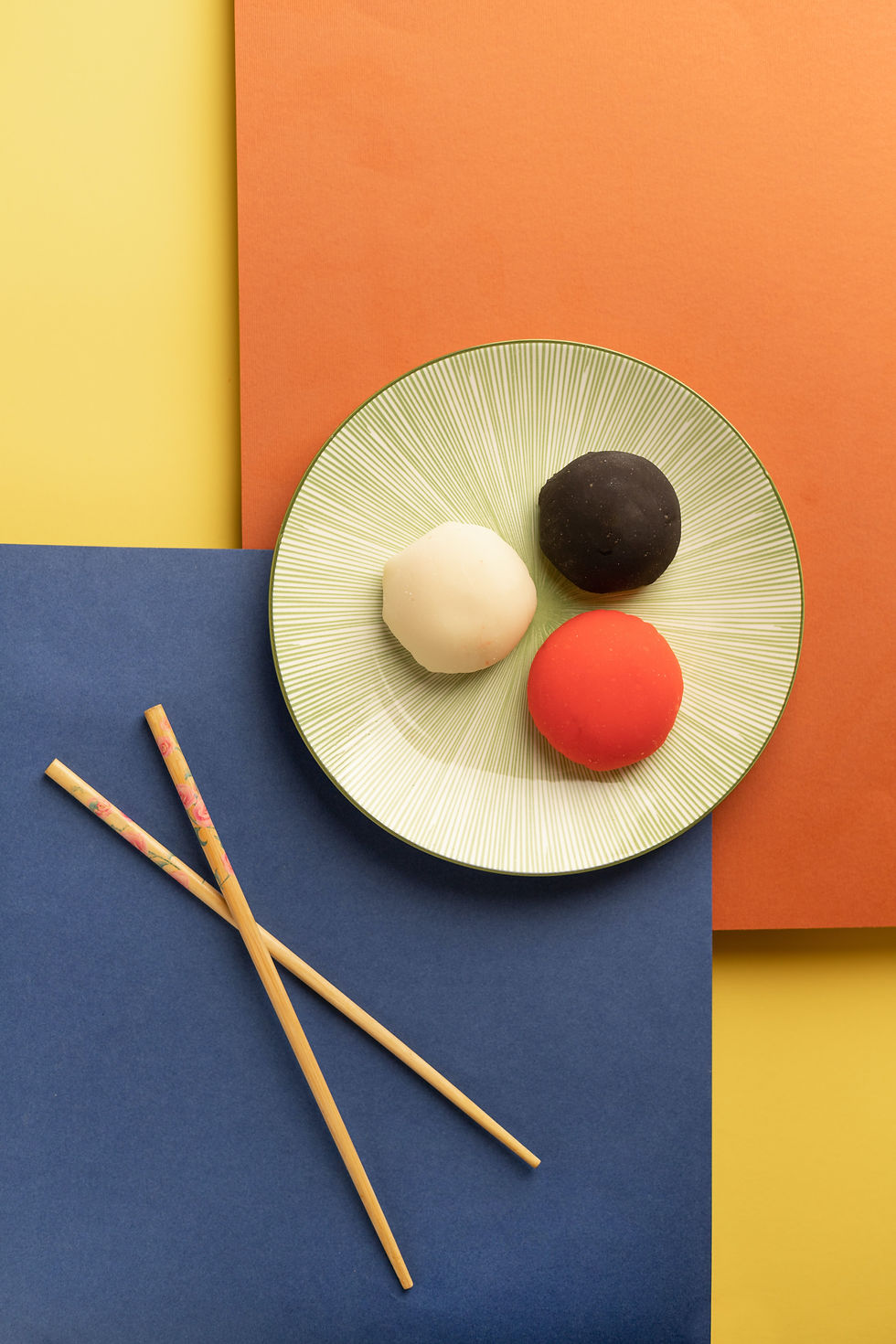 Plate with three mochi (white, black, red) on a green pattern plate, set against orange, blue, and yellow background. Chopsticks nearby.