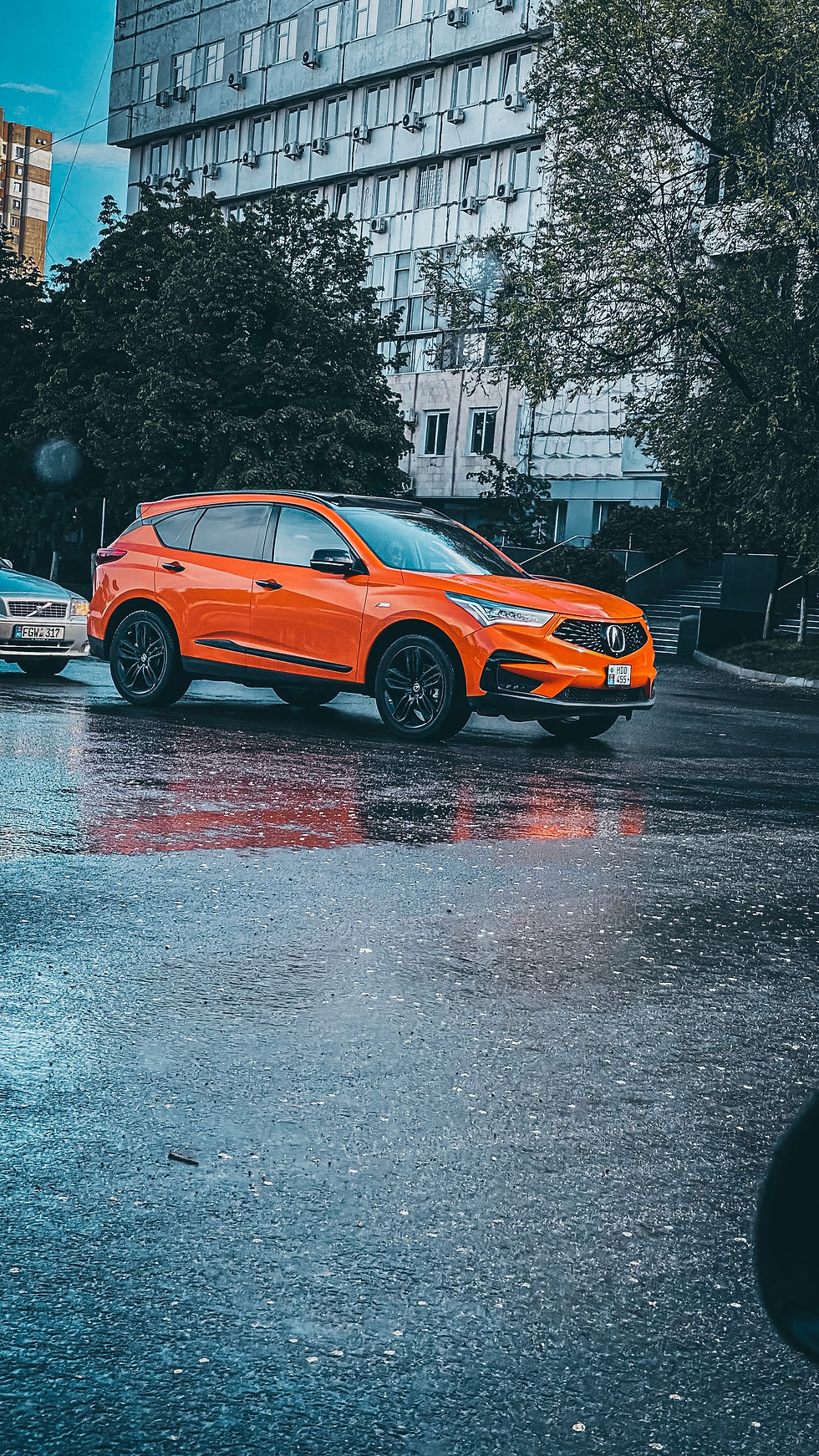 Orange SUV on wet road beside buildings and trees. Overcast mood with vehicle reflections on the pavement. Plates visible, no text.