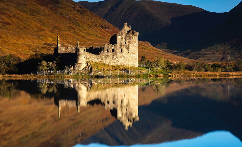 Ancient Urquhart castle ruins beside a calm lake, reflecting in the water. Autumnal hills in the background, under a clear blue sky.