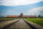 Railway tracks lead to a brick building with a central arch under a clear blue sky. Grass on either side creates a solemn atmosphere.