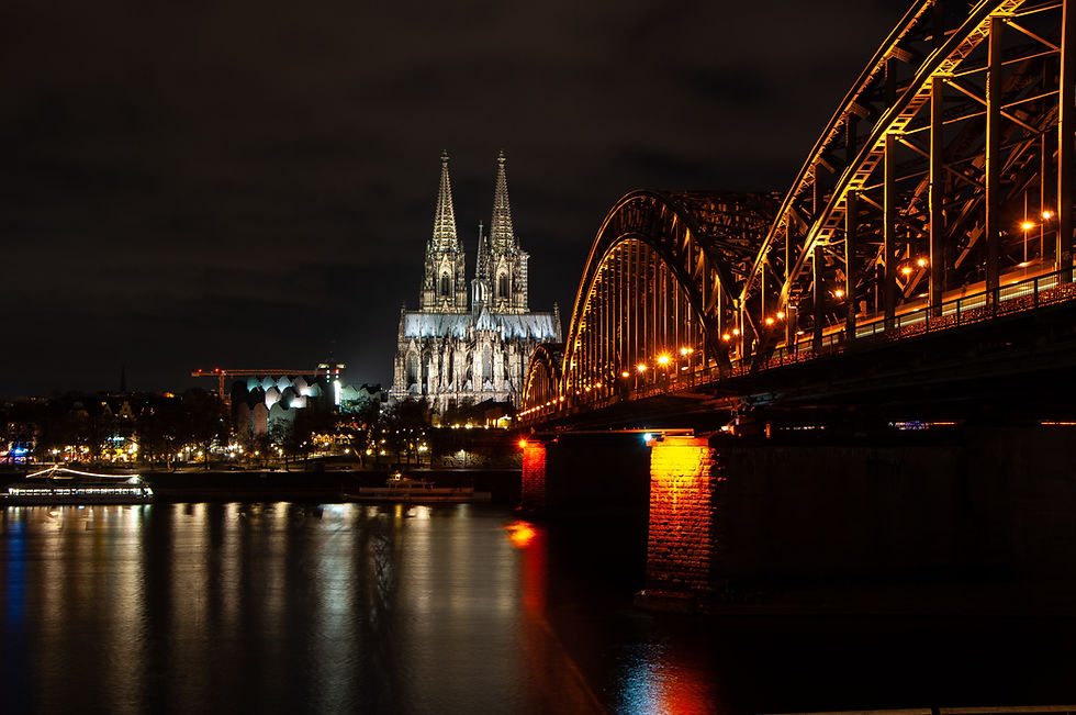 Illuminated Gothic cathedral and arched bridge at night with glowing lights. Reflective water and dark sky create a serene atmosphere.