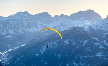 Paraglider with a yellow canopy soars over snow-covered mountains, creating a serene scene against a clear sky.