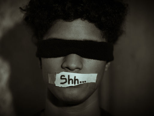 Blindfolded man with taped mouth showing "Shh..." in dim light. Intense, mysterious mood. Brown and black tones dominate. Symbolizes men being quieted due to impact of "toxic masculinity" narrative.