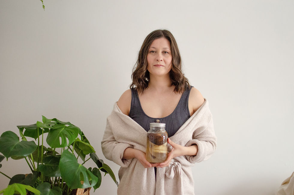 Woman in beige robe holding a jar with liquid and brown objects, standing beside a leafy plant against a plain background. Looks calm.