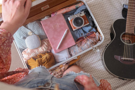 Open suitcase packed with clothes, a pink notebook, camera, and sunglasses beside a black guitar on a checkered bed.