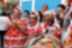 Three ladies dressed in traditional red dresses and hats play forro music with drums and a triangle