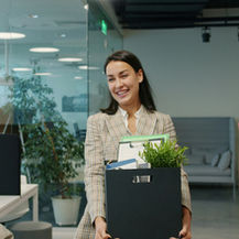 Smiling woman carrying a box with a plant and files in a modern office, symbolizing the excitement of changing career, while a man works at his desk nearby.