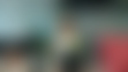 Smiling woman carrying a box with a plant and files in a modern office, symbolizing the excitement of changing career, while a man works at his desk nearby.