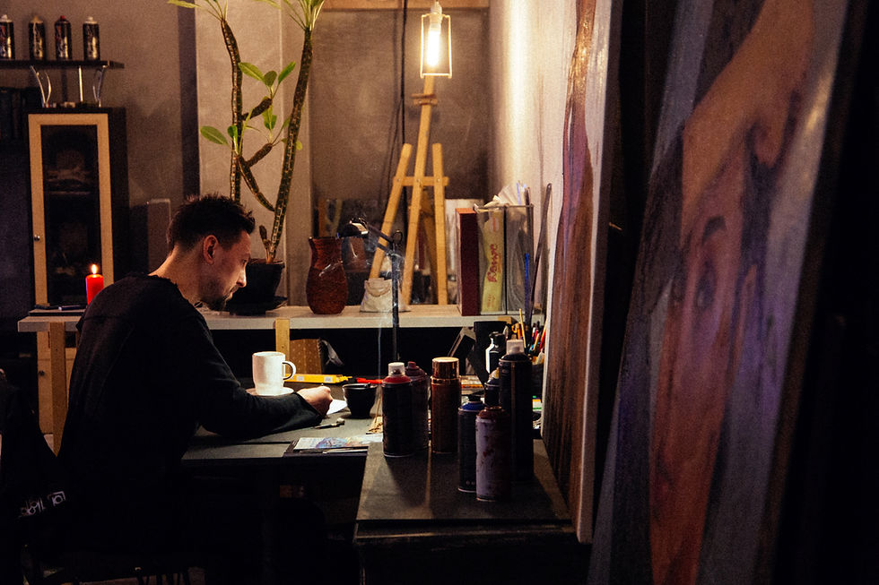 Artist at a cluttered desk, drawing under warm light. Paint cans, a red candle, and art supplies surround him. Canvas with portrait visible.