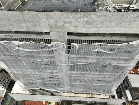 Aerial view of a concrete building under construction, covered with protective white netting. The rooftop and surrounding cityscape are visible.