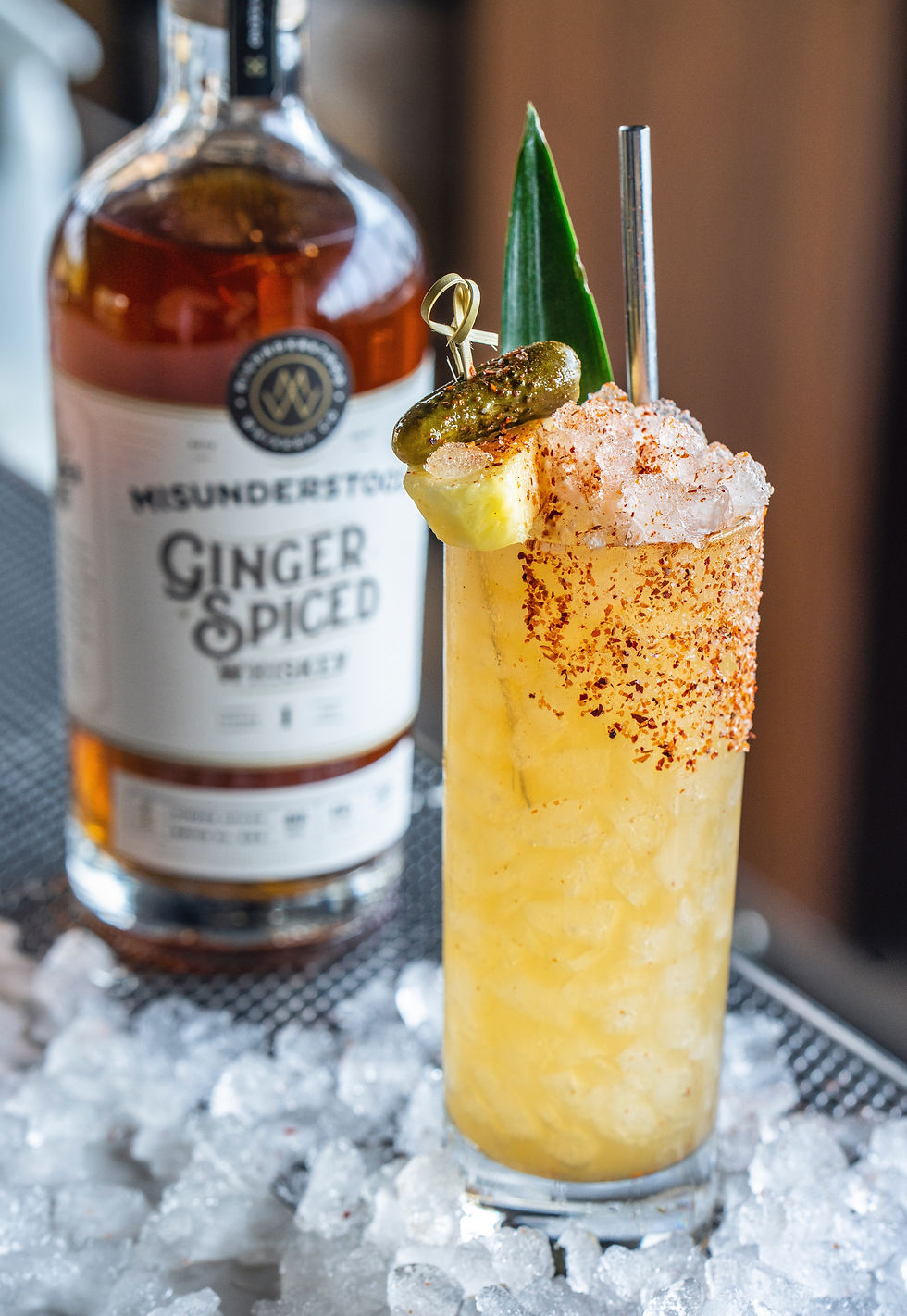 Icy ginger cocktail garnished with pineapple, jalapeño, and chili powder, beside a ginger spiced whiskey bottle on a metal surface.