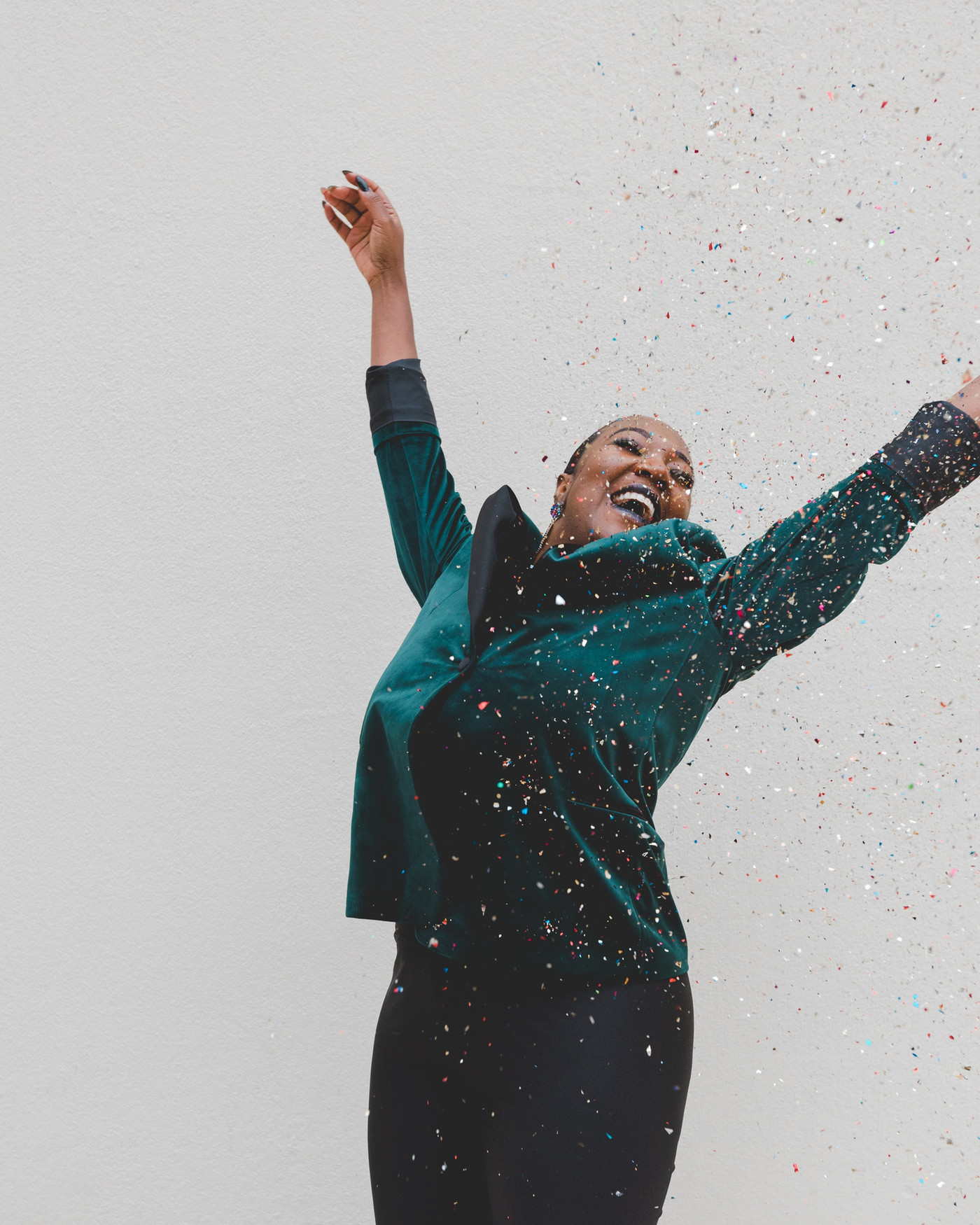 Smiling woman in teal top and black pants throws up her arms as colorful confetti falls against a white background.