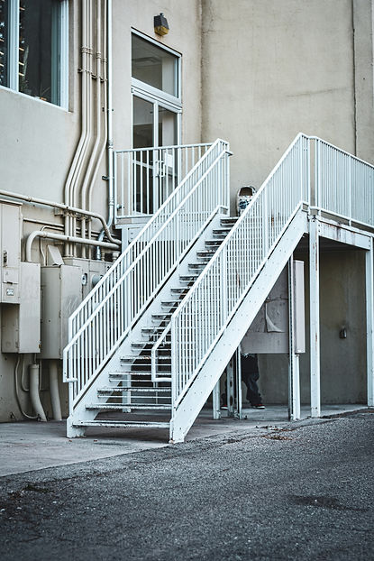 A white metal staircase ascends alongside an industrial building, with pipes and utility boxes visible