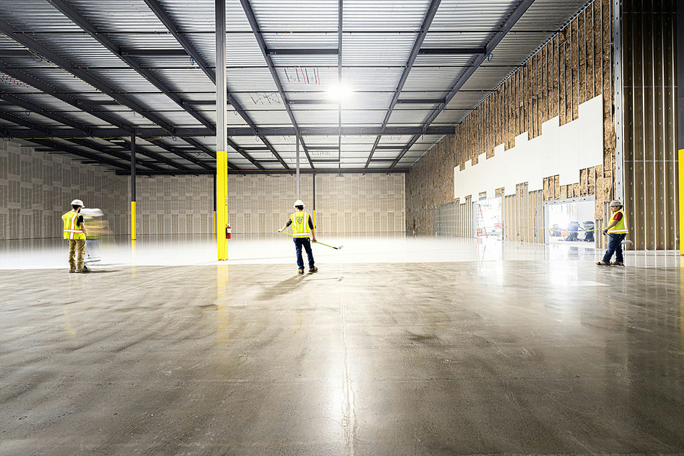 Workers in yellow vests and helmets polish a concrete floor in a large, bright warehouse with metal beams and unfinished walls auditing lights.