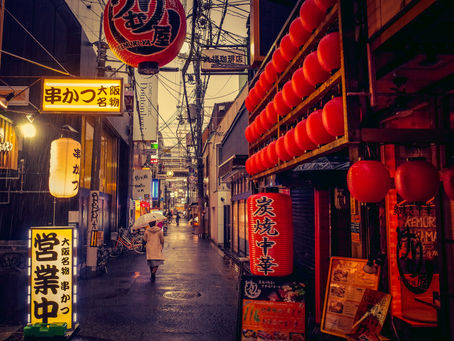 A person with an umbrella walks through a narrow street lined with red lanterns and yellow signs surrounding thriving Japanese businesses.