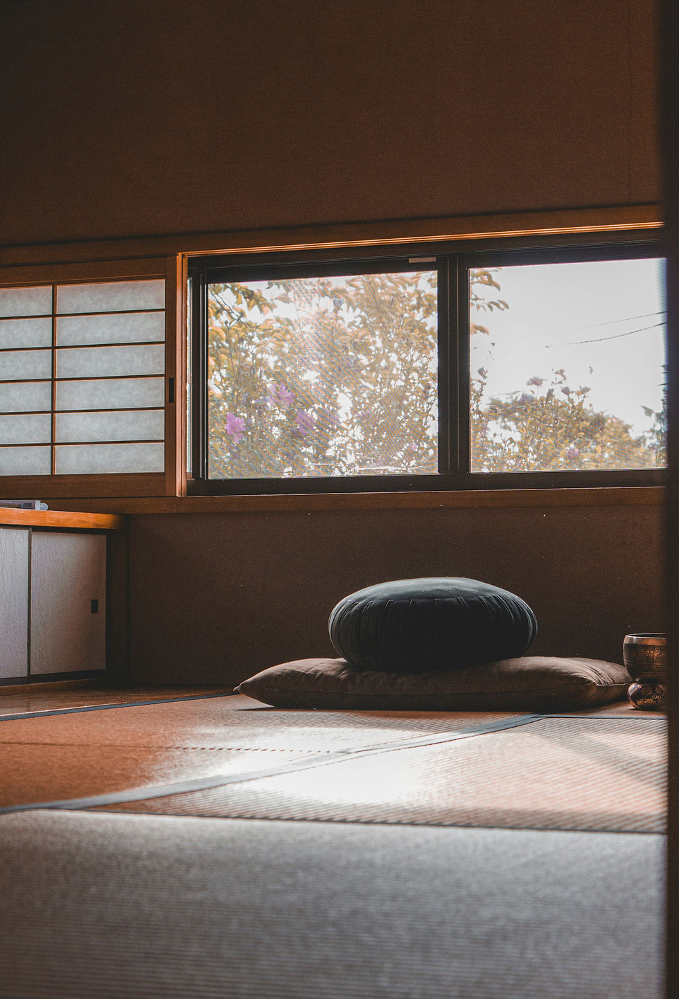 Quiet tatami room with stacked cushions by a sunlit window, overlooking trees and purple flowers, warm and serene atmosphere