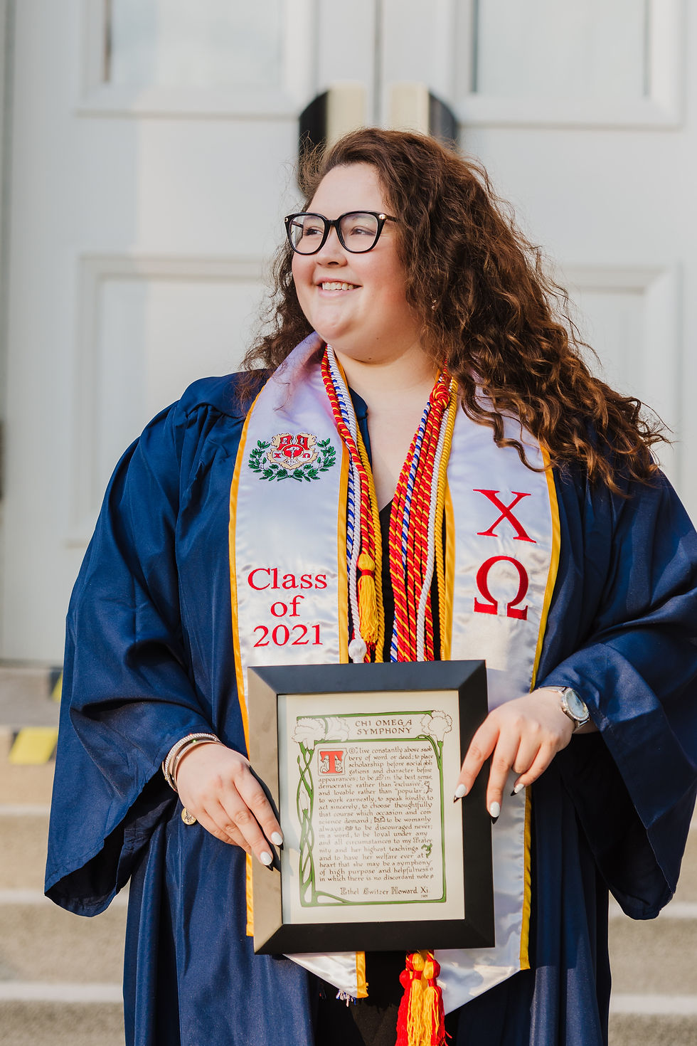 A woman with long curly hair poses at a university graduation event with her diploma or an award. She is wearing university robes in addition to tassels and a sash.