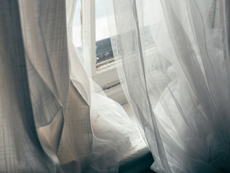 Open book on white table with sunlight, vase with greenery, and stacked dice. Sheer curtains create a serene, calm mood.