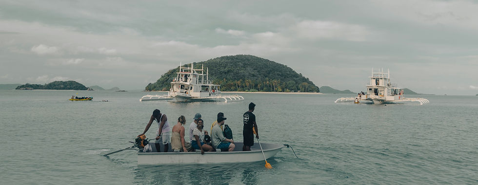 A group on a small canoe or boat being paddled across the ocean while two larger ships are seen in the background, along with two small islands.