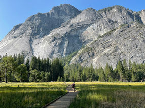 A cyclist rides on a wooden path through a green meadow, with towering gray mountains and a clear blue sky in the background.