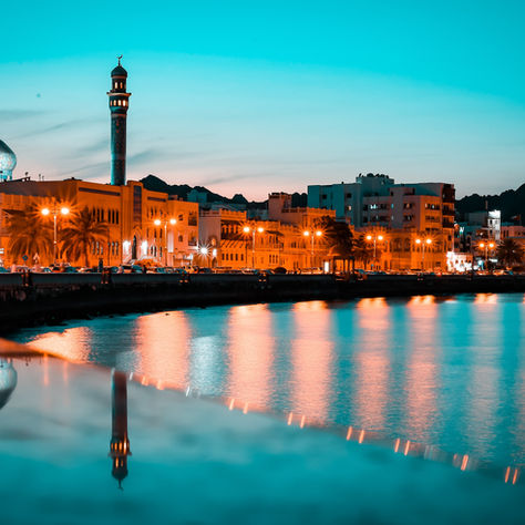 Beautiful evening view of Muscat's waterfront with mosque minaret, illuminated buildings, and calm reflections on the water under a vibrant blue sky.