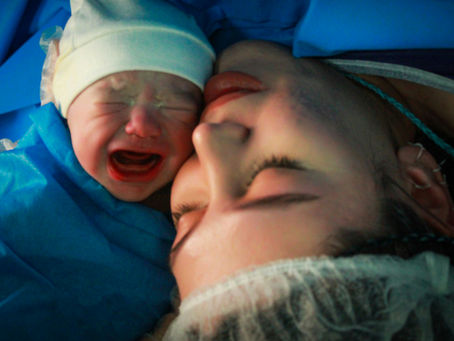 Newborn baby crying next to a woman's face, both lying under a blue surgical cloth. The woman appears calm, wearing a medical cap.