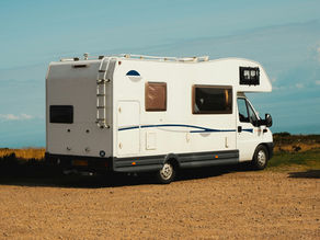 White RV parked on a dirt road with grassy fields and a blue sky in the background. License plate: "DK05 FSY." Relaxed, scenic vibe.