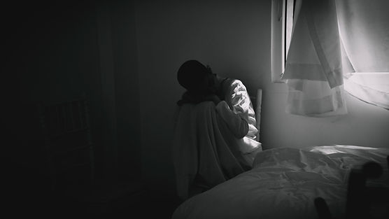 A person in a white outfit sits curled up in a dim room, looking somber. Light streams in from a window with curtains, beside a bed.