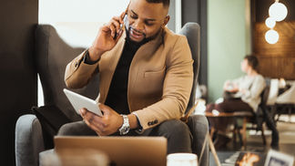 A prospective traveler talks on his phone while glancing at a tablet in a warmly lit café lounge, an open laptop nearby, mid due-diligence call.