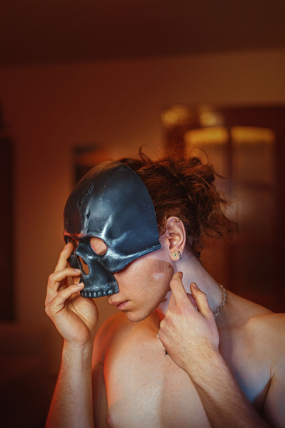 Person with red hair holds a black skull mask to their face in a warm-lit room. They wear an earring and a chain necklace, creating a mysterious mood.
