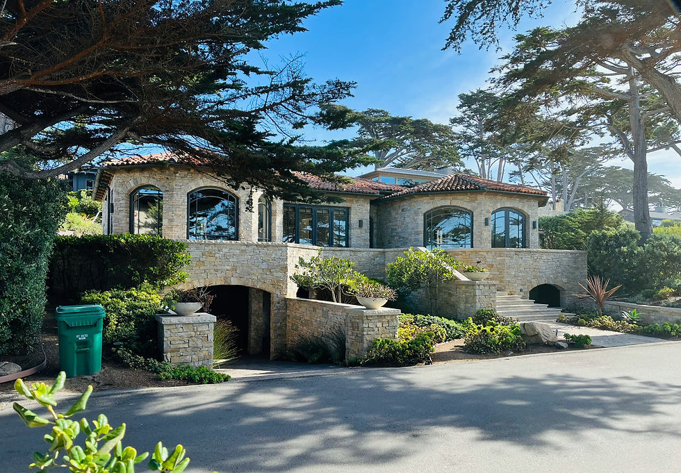Stone house with arched windows and red tile roof, surrounded by lush greenery and trees under a clear blue sky. Green bin in front.