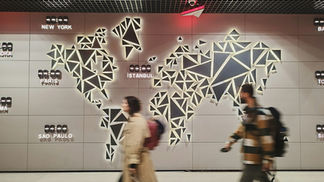Two travelers with backpacks and rolling suitcases walk past an illuminated geometric world map on an airport wall, with world-city clocks for New York, Paris, Sao Paulo, Istanbul, Rome, and Tokyo glowing beside it