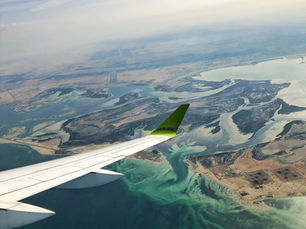 Airplane wing above a coastal landscape with turquoise waters and sandy beaches. The wingtip reads "airBaltic." Sky is partly cloudy.