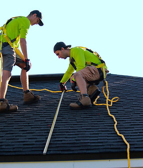 Two roofers in safety harnesses installing shingles on a steep residential roof