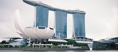 The Marina Bay Sands hotel with its three tall towers connected by a rooftop platform, and the lotus-shaped ArtScience Museum in Singapore, viewed from across the water on a cloudy day.