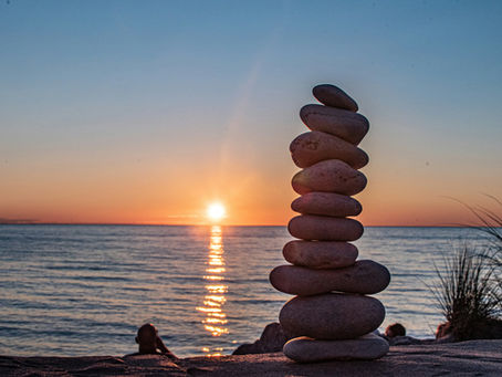 Stacked stones on a beach at sunset, with silhouettes of people watching the calm sea and orange sun.