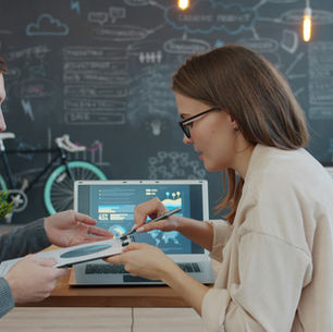 Two coworkers review charts and a 45% dashboard on a laptop in a modern office, with a chalkboard wall and bicycle behind them.