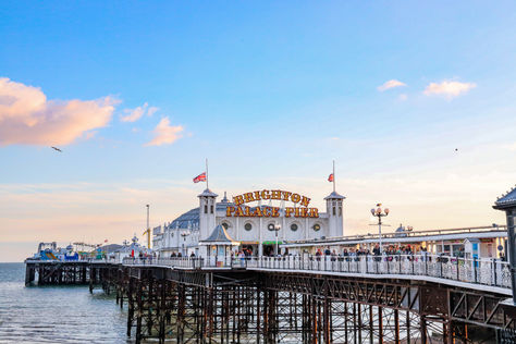Brighton Palace Pier stretches over the sea under a blue sky. Visitors walk along the pier, with flags and a sign visible. Peaceful ambiance.