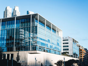 Modern glass building reflecting nearby structures under a clear blue sky, with minimal graffiti on lower walls and trees in the foreground.