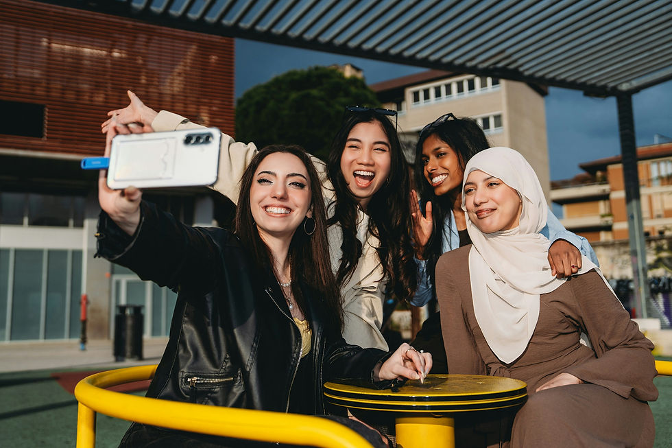 Four smiling women pose for a selfie at an outdoor campus courtyard with modern buildings and yellow tables.