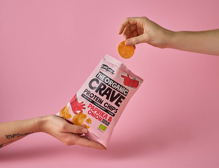 Two hands against a pink background. One holds a bag of "The Organic Crave" paprika & onion chips, while the other takes a chip. Located at commercial product video studio.