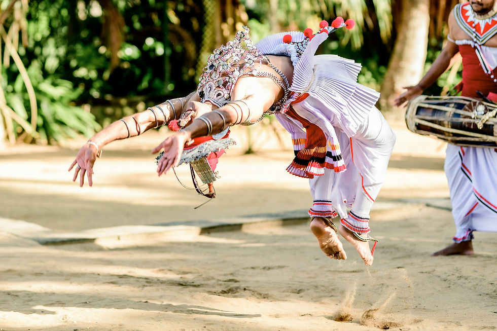 Photo Credit: Dinuka De Silva
A dancer in ornate costume performs a leap on sandy ground, with a musician playing a drum in the background. Vivid reds and whites.