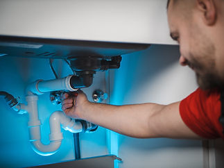 Man in red shirt fixing plumbing under sink, illuminated by blue light